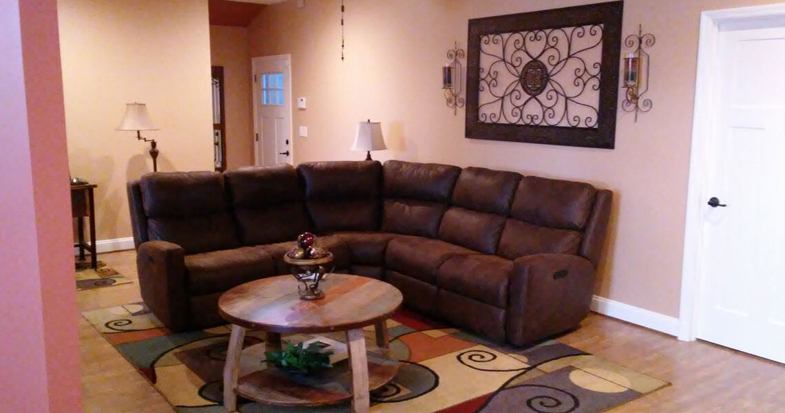A cozy living room with a brown sectional sofa, a round wooden coffee table on a colorful rug, and a decorative wall piece above the couch. Two lamps are in the background near a white door.