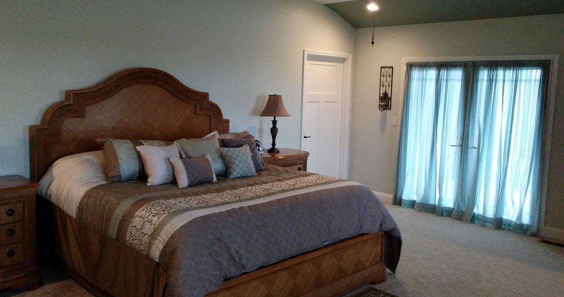 A cozy bedroom with a large wooden bed, adorned with patterned bedding and several pillows. Two matching wooden nightstands flank the bed, each with a lamp. Sheer curtains cover a glass door, and the walls and ceiling are painted in soft, neutral tones.