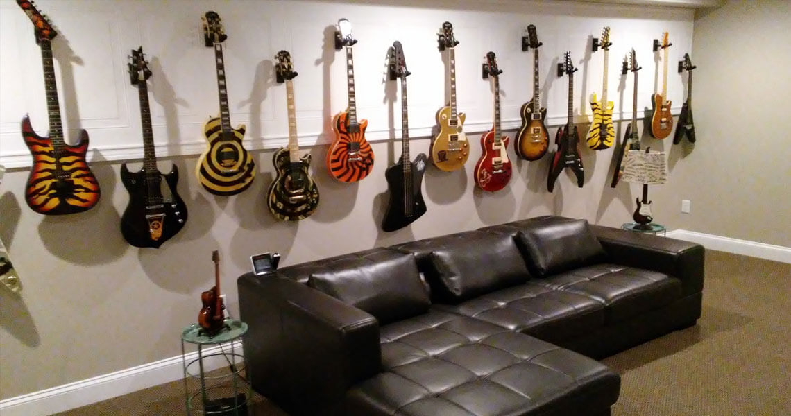 A collection of electric guitars with various colorful designs, including tiger stripes and sunbursts, is mounted on a wall. In front of them is a black leather sectional sofa with small glass tables on each side.