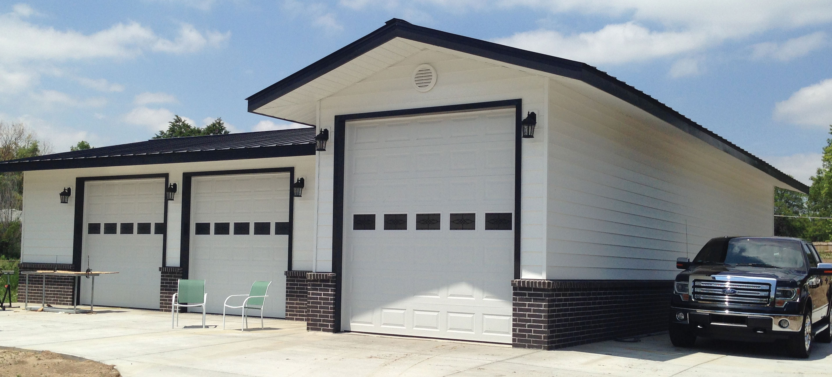 A modern garage with three large white doors and black trim offers more than just storage solutions. Two green chairs invite conversations, while a black pickup truck is parked beside it, ready for the next adventure under a partly cloudy sky that seems to ask its own questions.