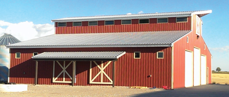 A large red barn with a corrugated metal roof stands proudly under the clear blue sky, showcasing the company’s expert construction. Four small windows adorn its upper section, while closed barn doors complete its sturdy facade. It sits amidst flat golden fields, earning kudos for its timeless charm.
