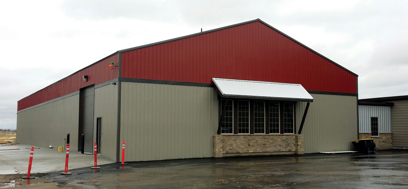 A large industrial building with a red and gray metal exterior, crafted by a reputable construction company, stands under an overcast sky. It features a sloped roof, large windows, and a stone accent at the base. Tall orange traffic cones are neatly lined up near the structure.