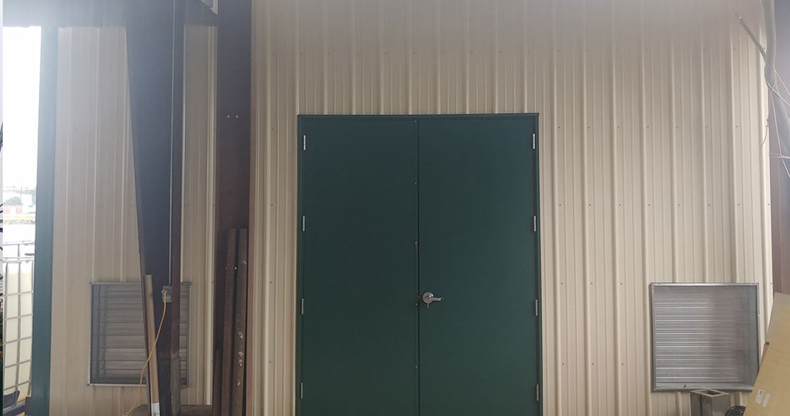 A green metal double door set in a beige corrugated metal wall, flanked by two small square vents. The building exterior appears industrial. The ground is concrete, with some equipment partially visible on the sides.