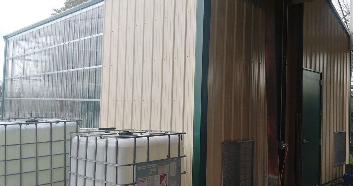 A beige metal building with a partially transparent roof extension. Large white plastic containers are placed on the ground outside. A green door is on the side of the building.
