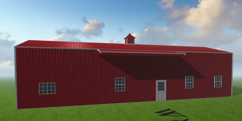 A large red barndominium with a white door and windows stands against a grassy field as the sunset casts a warm glow across the sky. The barn has a pitched roof and a small cupola on top.