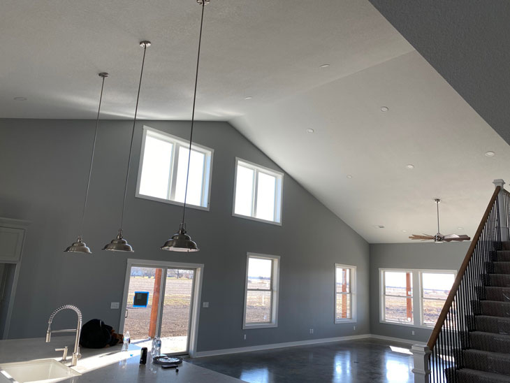 Spacious open-concept living area with high ceilings, large windows, and modern pendant lighting. Stairs on the right lead to an upper level. Natural light fills the room, highlighting the minimalist design and neutral color palette.