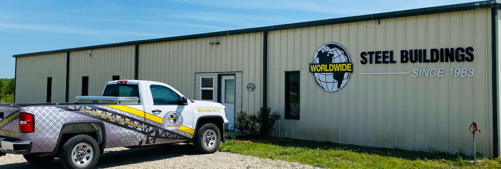 A white pickup truck with company branding is parked outside a beige industrial building. The building displays a sign that reads "WORLDWIDE STEEL BUILDINGS SINCE 1983" with a globe logo. It's a sunny day.