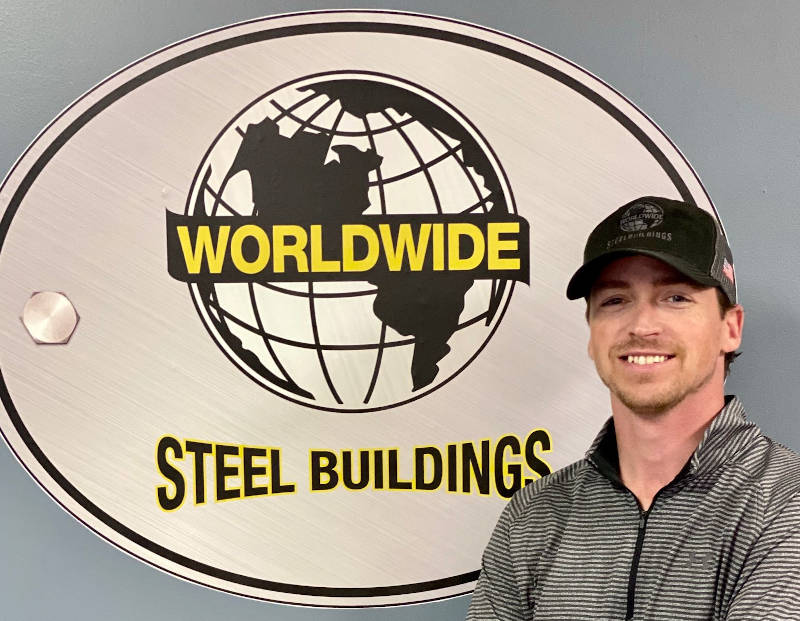A person wearing a dark hat and gray jacket stands smiling in front of a large sign that reads "Worldwide Steel Buildings," featuring a globe graphic. The backdrop, with its neutral-colored wall, highlights the prominence of steel building craftsmanship.