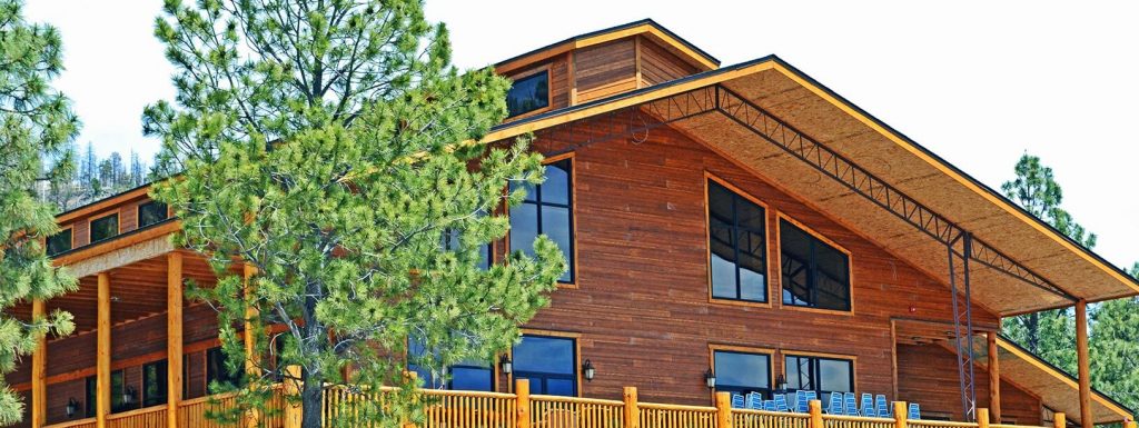 A large wooden cabin with modern architectural design is surrounded by tall pine trees. The structure, boasting expansive windows and a spacious deck, seamlessly blends rustic charm with contemporary style and comes with a dependable steel building warranty for peace of mind.