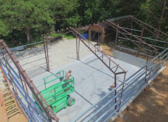 From above, the construction site on College Hill reveals a partially built metal frame structure. A worker on a green lift navigates inside, amidst trees, gravel, and equipment. This framework outlines a future building atop its concrete foundation—perhaps a new shop in the making.