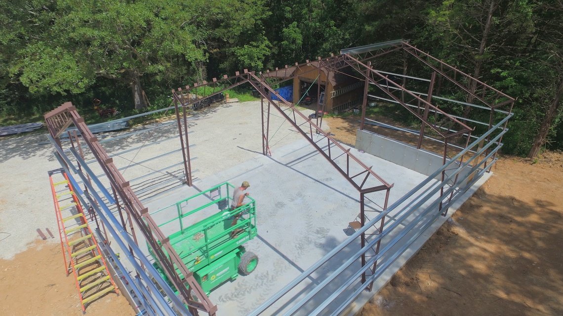 From above, the construction site on College Hill reveals a partially built metal frame structure. A worker on a green lift navigates inside, amidst trees, gravel, and equipment. This framework outlines a future building atop its concrete foundation—perhaps a new shop in the making.