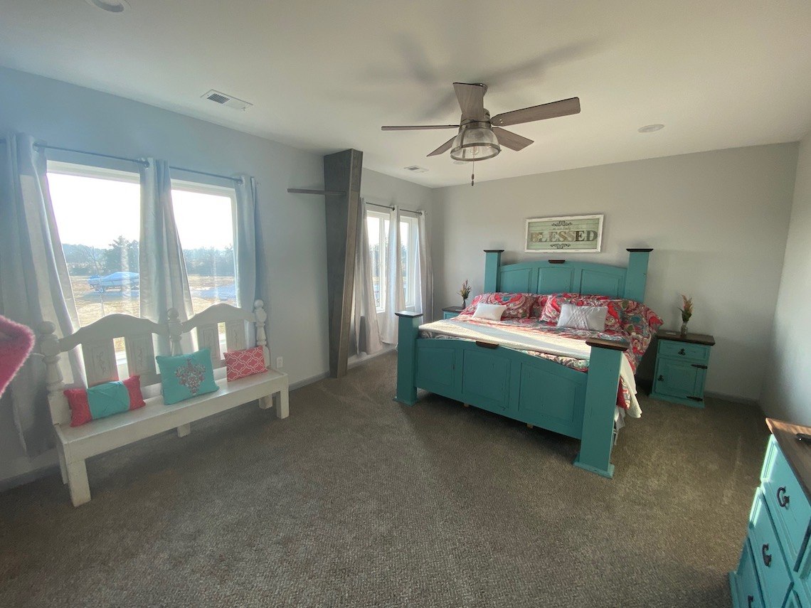 A bright country bedroom with a large turquoise bed, floral bedding, and a matching nightstand. A white bench with colorful pillows sits by the windows, covered in light curtains, perfect for enjoying the barndo's serene views. A ceiling fan hangs elegantly from the ceiling.