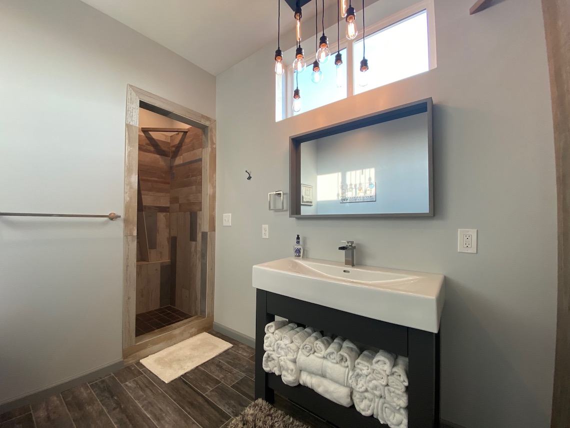 This country-inspired bathroom features a modern vanity with a wide sink and rolled towels neatly on a shelf beneath. A large mirror is mounted above, while the walk-in shower boasts wood-like tiles. Hanging light bulbs and natural window light complete this barndo aesthetic haven.