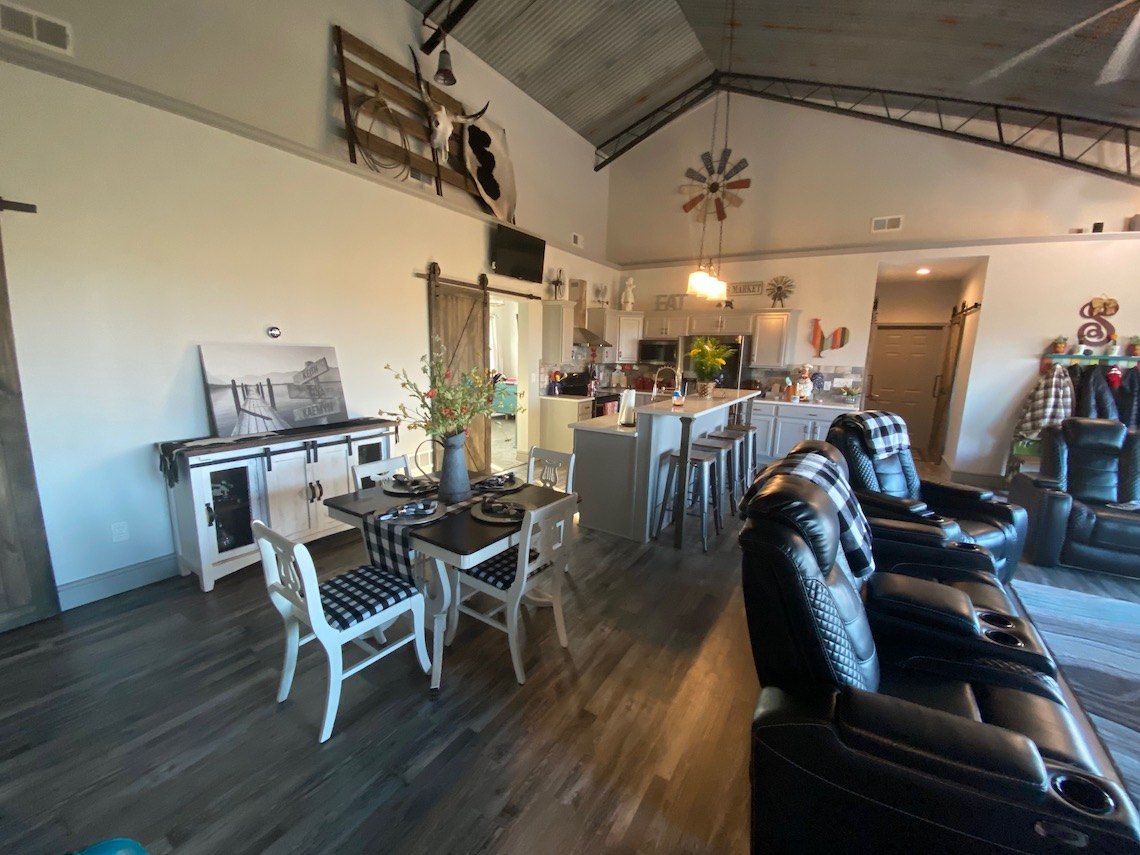 A cozy, rustic living and dining area in a barndo style features black leather recliners, a dining table with checked placemats, and country farmhouse decor. The room includes wooden floors, a kitchen island, and homey decorations like wreaths and large wall art.
