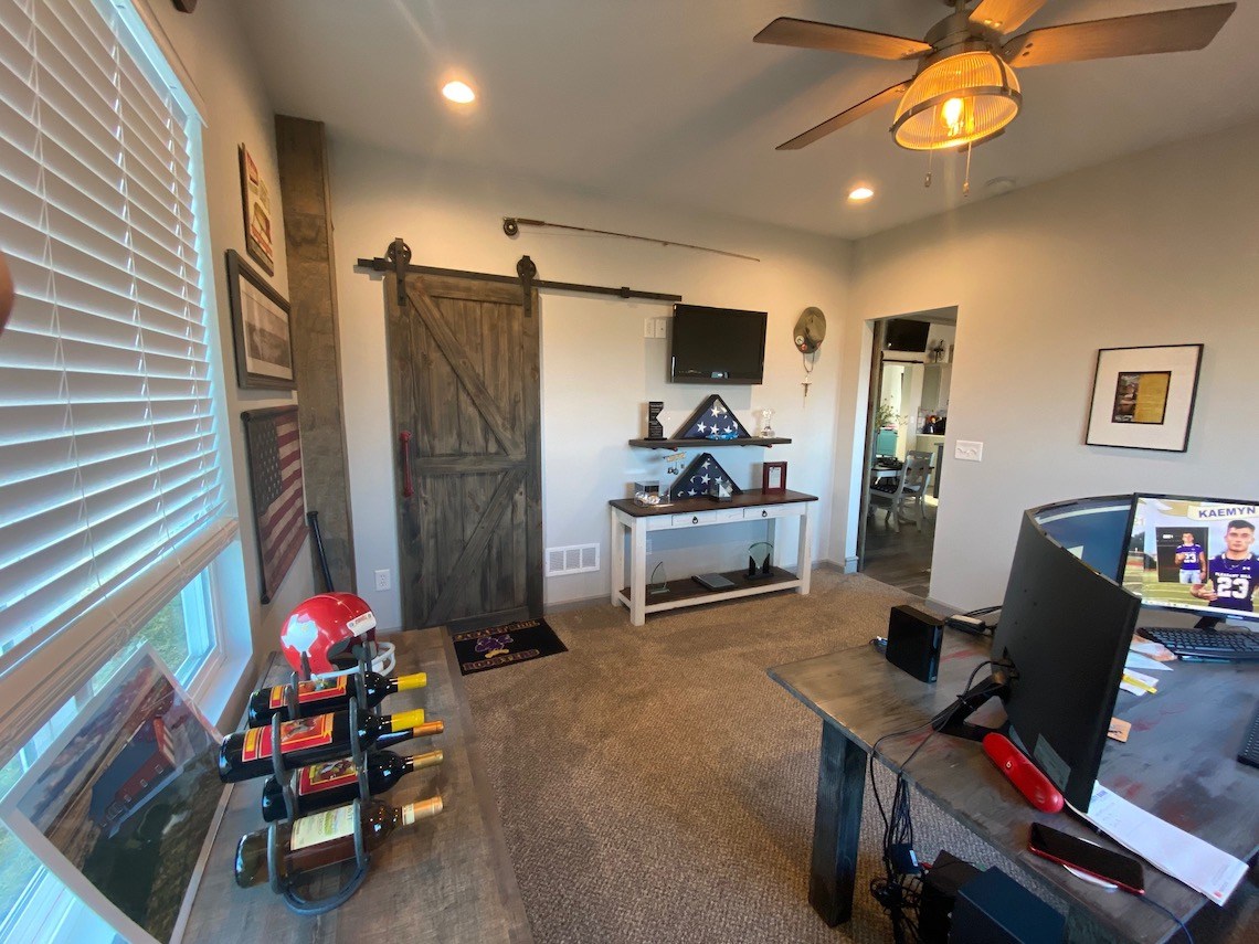 A cozy home office with a computer on a wooden desk, a wall-mounted TV, and a barn door for that country charm. Shelves display folded flags and memorabilia. A ceiling fan whirs above, framed pictures line the walls, and natural light streams through the window on the left.
