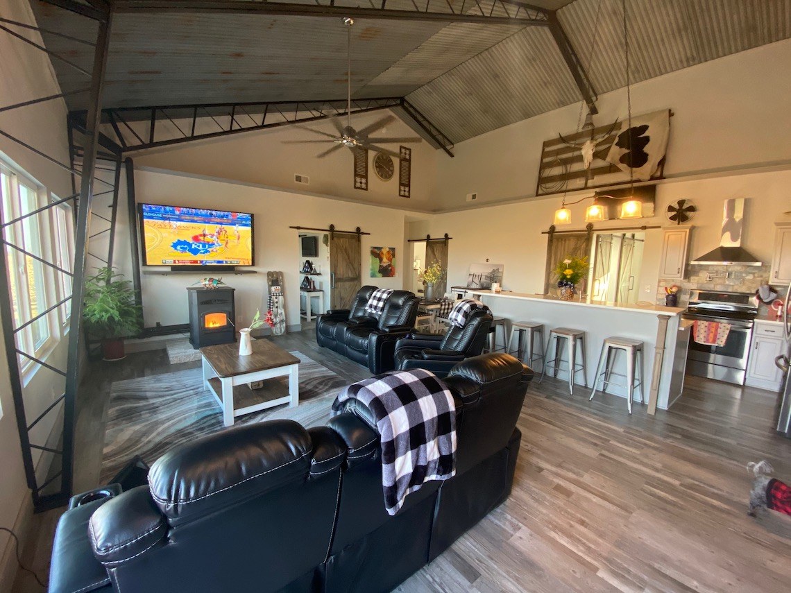 A spacious living room with vaulted ceilings, featuring leather sofas, a coffee table, and a wall-mounted TV showing a basketball game. This barndo-style space combines modern rustic decor with country charm, boasting an open kitchen with bar stools at the counter, plants, and wall art.