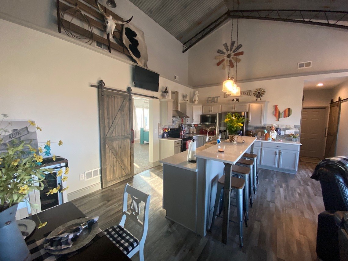 A cozy, rustic kitchen and dining area with a high ceiling embodies the charm of a country barndo. Features include a wooden island with bar stools, farmhouse decor, sliding barn doors, and various plants. The space has a warm, inviting atmosphere with natural light filtering in.