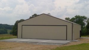 A large beige metal barn, resembling a hangar, with a closed front door is situated in a rural area near Warsaw. The barn sits on a concrete foundation surrounded by grass and trees under a cloudy sky.
