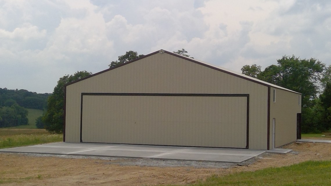 A large beige metal barn, resembling a hangar, with a closed front door is situated in a rural area near Warsaw. The barn sits on a concrete foundation surrounded by grass and trees under a cloudy sky.