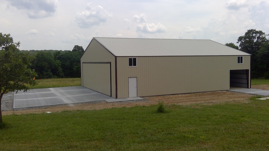 A large beige metal hangar with a sloped roof rests on a concrete pad near Warsaw. It features a spacious sliding door and a smaller entrance, surrounded by grass and trees beneath a cloudy sky.