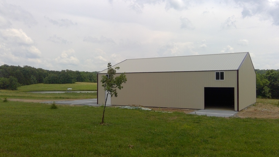 A large beige barn with a gray roof stands in an open field near Warsaw, with a few small trees dotting the landscape. The cloudy sky looms above, and in the background, a small pond is surrounded by greenery and rolling hills resembling an idyllic scene from an old-world hangar setting.