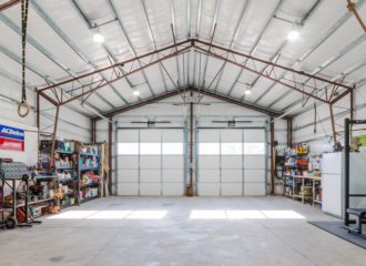 A spacious garage with high ceilings and metal beams, akin to an Arizona workshop, featuring two large garage doors. The interior includes shelves stocked with tools and equipment, a red tool chest, exercise gear, and gymnastic rings hanging from the ceiling.