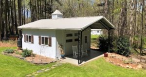 A small white metal building with a peaked roof sits cozily in a grassy area surrounded by Carolina trees. It features a covered porch with two rocking chairs, inviting relaxation. The area around the workshop is thoughtfully landscaped with shrubs and plants.