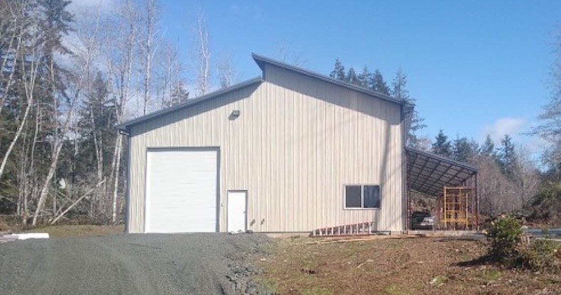 A light beige metal warehouse building in Olympia with a large garage door and a smaller side door stands surrounded by trees. A shop-like covered structure is attached to one side, and there's a stack of materials nearby. The sky is clear and sunny, evoking a dreamy atmosphere.