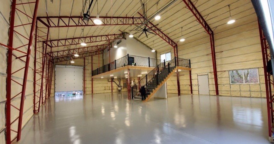 The interior of the Olympia warehouse feels like a dream space, with its high ceilings and red steel beams. Concrete flooring stretches beneath large garage doors, while a wooden mezzanine with railing and central staircase provides elevated shop space.