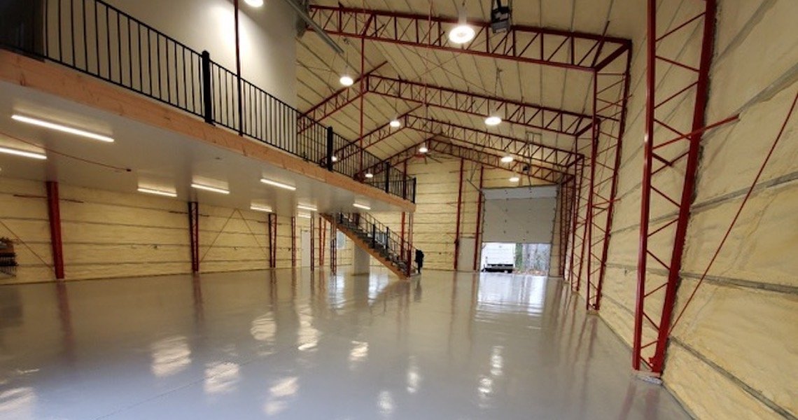 Spacious industrial warehouse interior with high ceilings, metal beams, and large overhead doors. This Olympia dream shop features a mezzanine level with railings and a staircase, and the floor is polished and clean. Natural light filters through the open door.