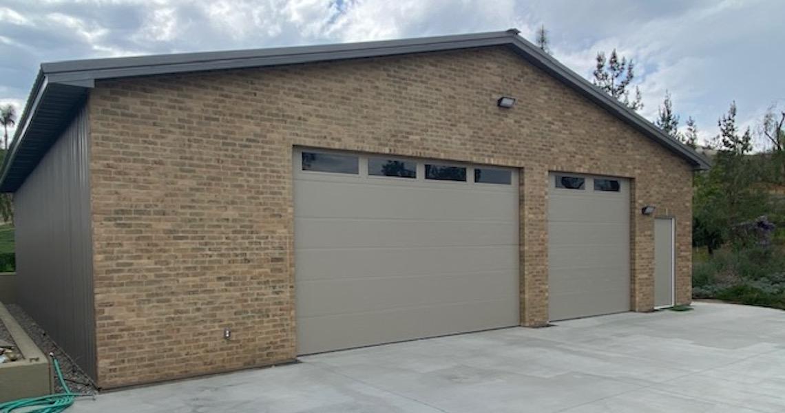 A large, single-story brick garage with two closed beige doors stands alongside the scenic Riverside. The building features a sloped roof and a small security light above. Surrounding it is a paved driveway framed by trees and shrubs in the background.