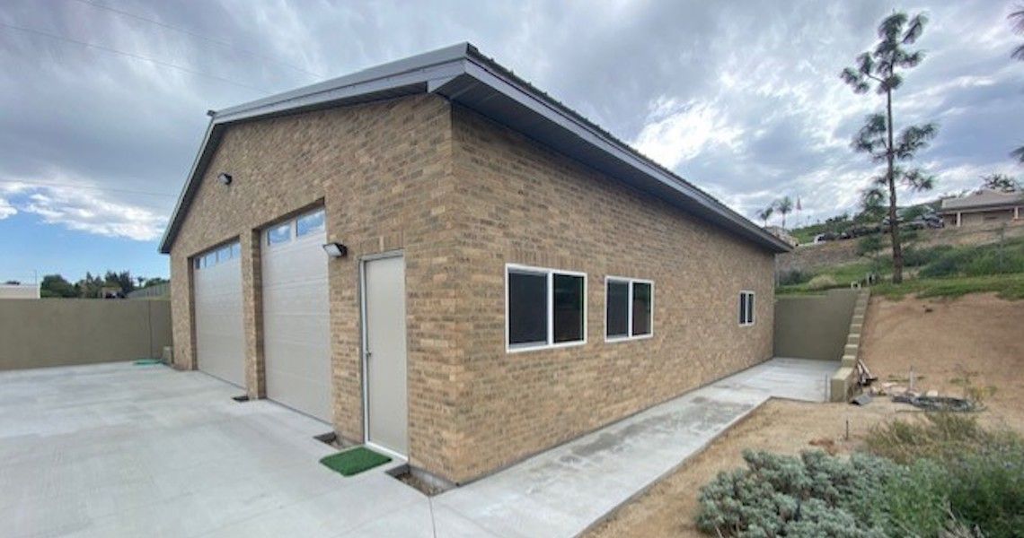 A large brick garage with two closed white doors and three windows stands on a paved area, nestled by a grassy hillside under a cloudy sky. The picturesque setting near the riverside adds charm to this functional space.