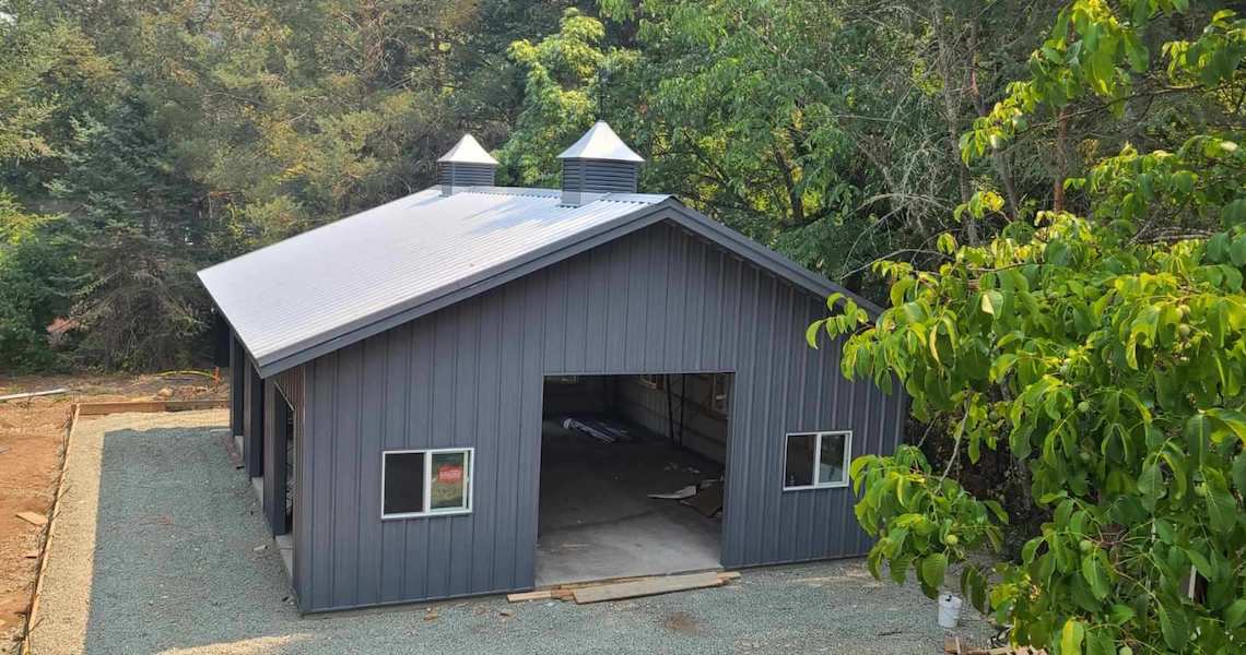 A dark gray metal building resembling a spacious gray garage with two small cupolas on the roof stands amidst the lush trees of St. Helena. It features a large open doorway, two windows, and rests on a gravel foundation.