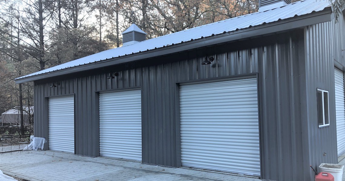 A gray garage in St. Helena, this metal building boasts three white roll-up doors on a paved lot surrounded by trees. Featuring a sturdy metal roof with two rooftop vents, it basks in natural light filtering through the wooded surroundings, offering a serene outdoor setting.