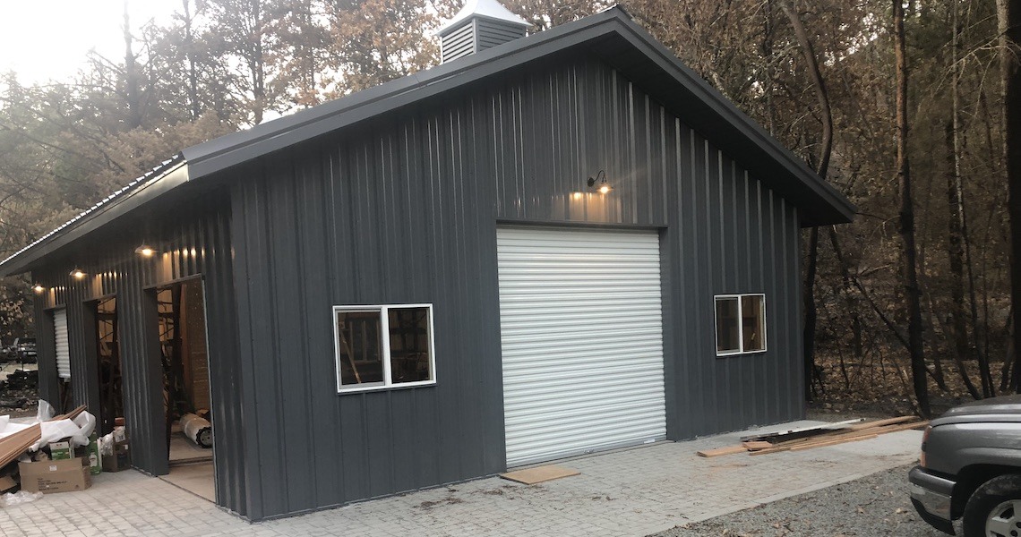 A gray metal garage with a St. Helena backdrop features a white roll-up door and two small windows, framed by surrounding trees. The paved ground hosts open boxes and construction materials to the left, creating a scene of subtle industrial charm.