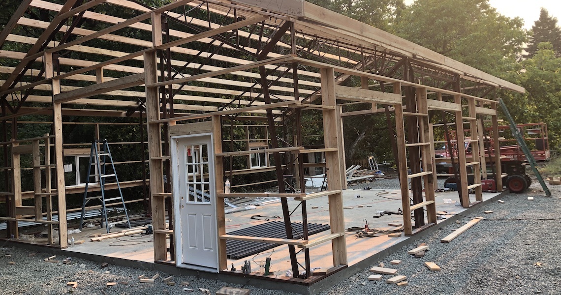 A partially constructed wooden barn with an open framework and a single white door stands proudly on a gravel and concrete base, reminiscent of St. Helena's rustic charm. Various construction materials and tools are scattered around, framed by trees in the background.