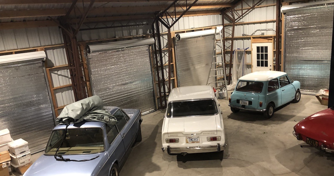 The Gray Garage houses four vintage cars, evoking nostalgia amid its classic decor. On the left, a blue car with a cover rests under the St. Helena sun streaming from above. Central stands a white car near a rolled-up door, while adjacent, the rear of a red car peeks beside a light blue compact.