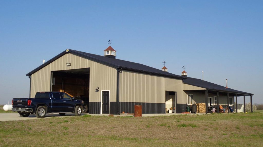 A large gray metal barn with a black roof and cupolas is situated on a grassy area under a clear blue sky. A black pickup truck is parked nearby, and the barn doors are open, revealing the interior. Logs and barrels are placed beside the barn.
