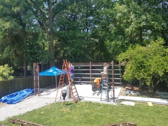 People are working on assembling a metal framework in a backyard surrounded by trees. They are using ladders for construction, with tools and materials scattered around. A blue canopy provides shade in the sunny setting.