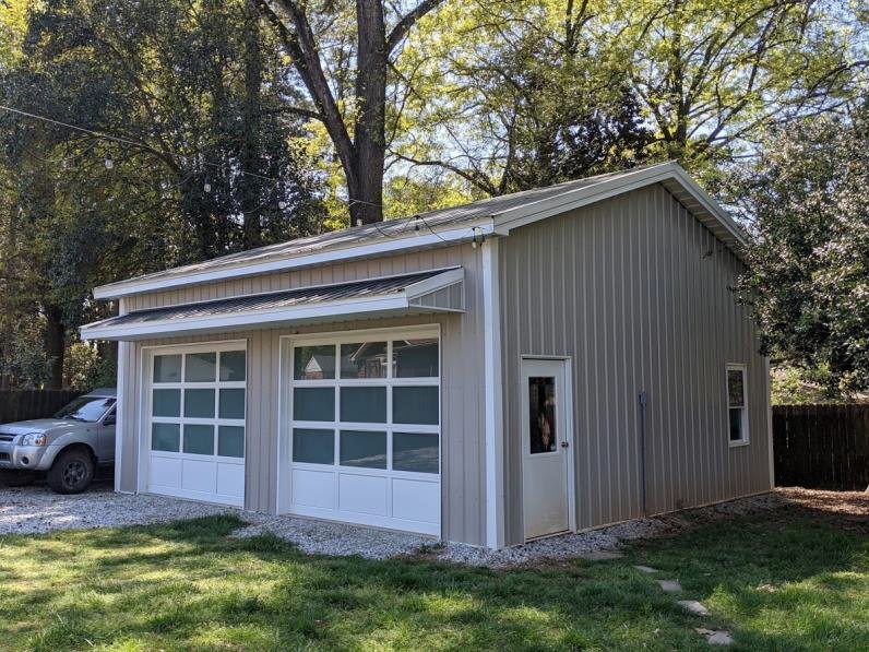 A metal garage with two large windows and a white door sits on a patch of gravel. A car is partially visible on the left. The garage is surrounded by greenery and trees under a clear sky.