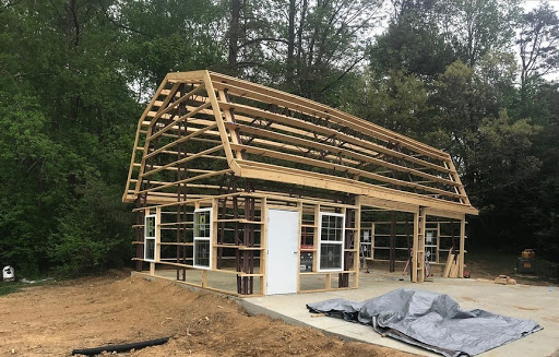 A half-built Gambrel barn stands with a wooden frame structure on a concrete foundation. The frame includes an A-frame roof and multiple window openings. In the backdrop, trees sway gently while a tarp lies on the ground nearby, marking progress by the Leino Family.