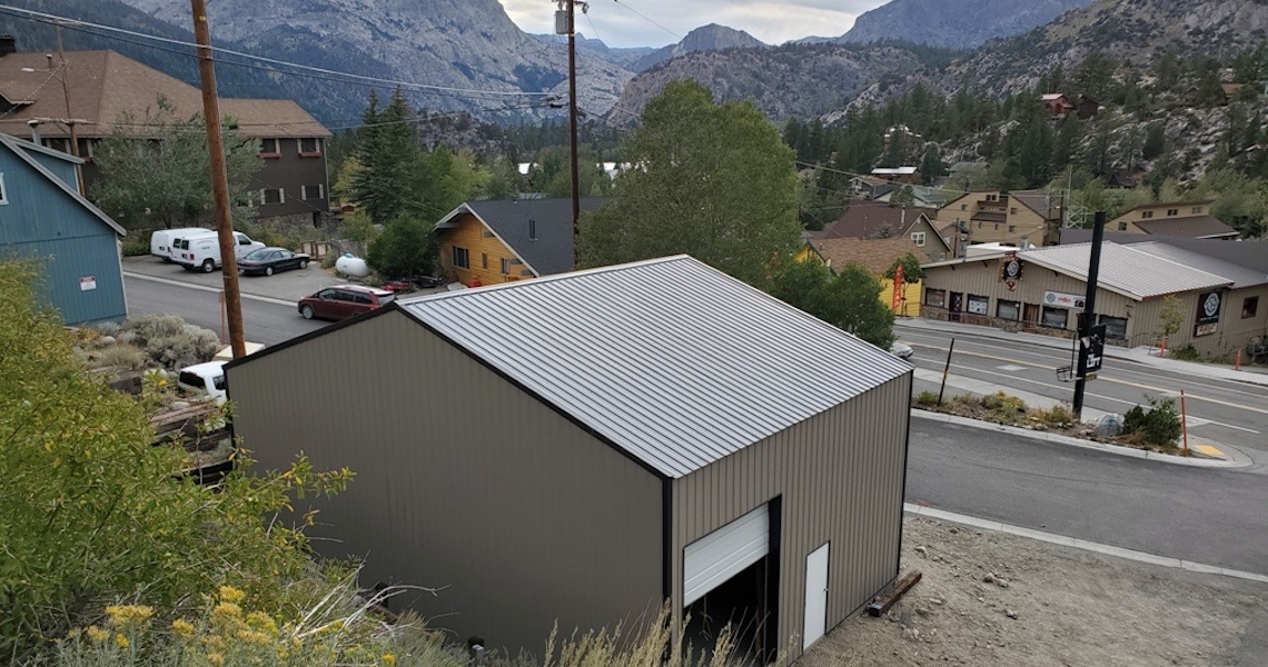 A small metal garage with a slanted roof is perched on a hillside overlooking a quaint California town. Several houses and parked vehicles are nearby, with mountains in the background under a cloudy sky. A road curves around the scene, enhancing its rustic charm.