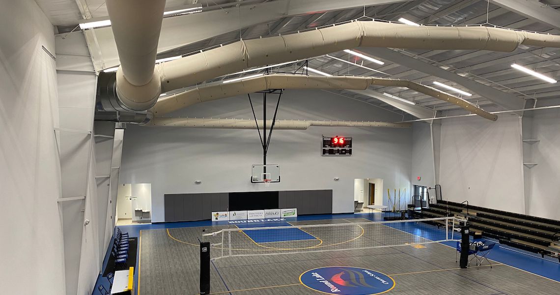 An indoor sports court at Round Lake Charter School features a basketball hoop and volleyball net, surrounded by bleachers. The ceiling hosts large ducts, while the scoreboard shows a score of 0-5. The floor is intricately marked for various sports activities.