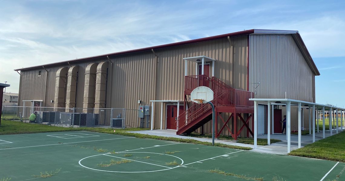 A large metal building with a red staircase stands next to an outdoor basketball court at Round Lake Charter School. The court's lines are clear, and a hoop with a white backboard awaits players. Grassy areas surround the scene under a partially cloudy sky.