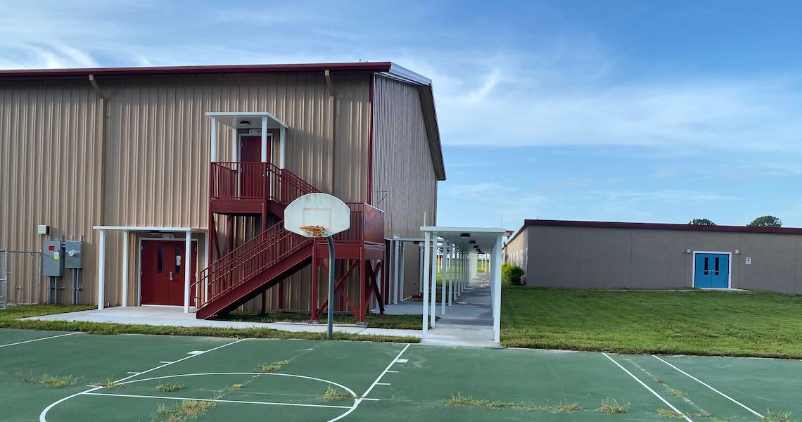 The Charter School near Round Lake features a school building with red exterior stairs, blue doors, and an adjacent walkway. Next to it lies a basketball court with an orange hoop. The sky above is clear and blue.
