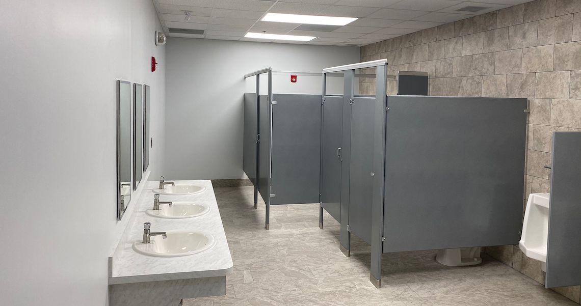 The public restroom at Round Lake Charter School features three sinks and mirrors on the left and three gray-stalled toilets on the right. The floor is tiled with a single urinal visible. Bright lighting enhances the light-colored walls, creating a clean and welcoming atmosphere.