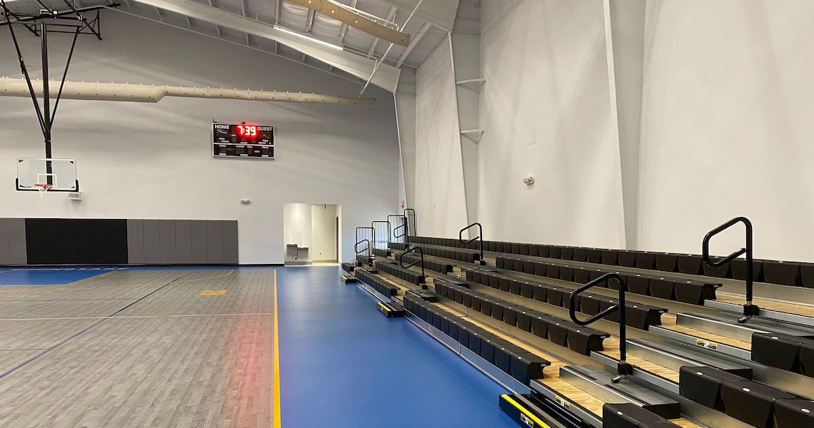 An empty indoor gymnasium at the Round Lake Charter School features bleachers on the right and a basketball hoop on the left. The digital scoreboard displays zeros, while the blue and gray floor is marked with game lines, ready for action.