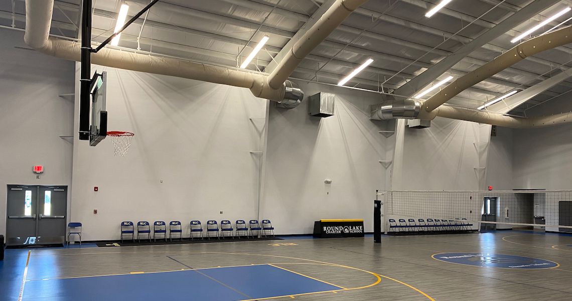 An indoor sports facility at Round Lake Charter School features a basketball court with a hoop on the left and a volleyball net on the right. Blue chairs are lined up against the wall, and there are ceiling lights and ventilation ducts above.