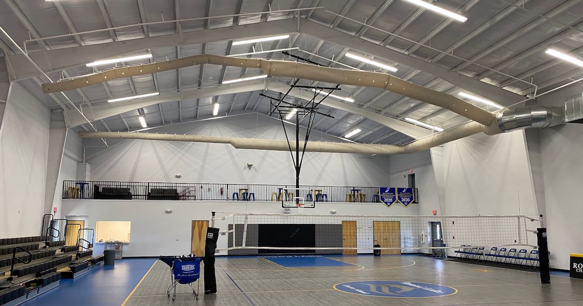 An indoor sports court at Round Lake Charter School boasts volleyball nets and a telescopic basketball hoop. Bleachers line the side, with banners proudly displayed on the upper railing. The ceiling features exposed piping and strategic lighting.