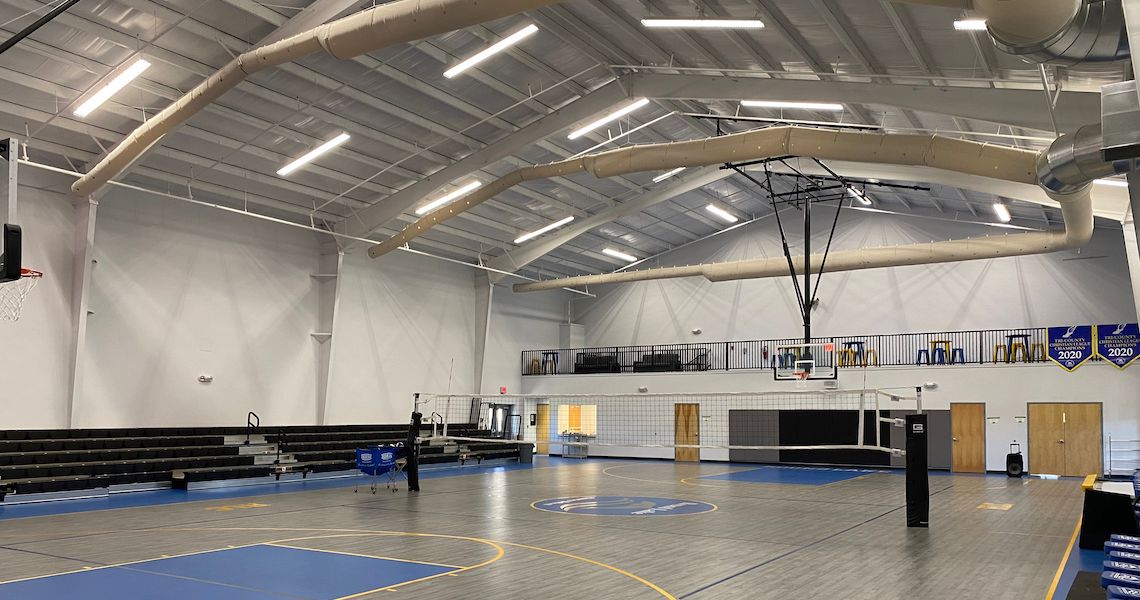 A modern gymnasium at the Round Lake Charter School features a basketball court with blue and gray flooring. On the left side, bleachers reside beneath exposed beams and ventilation pipes, while a volleyball net is set up on the court.